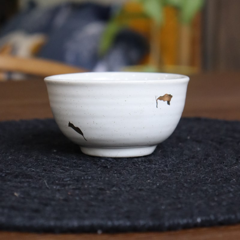 On a black place mat, the Ivory Ganga Small Bowl, a pure white and 24-karat gold ceramic small bowl you can buy online at Sukham Home, a sustainable furniture, gardening and home decor store in Kolkata, India