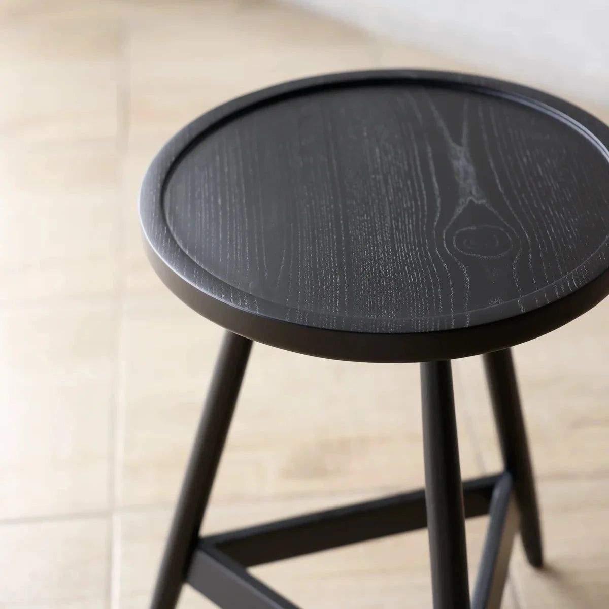 A round black wooden stool with a dark wood-grain finish and slim black legs, shown in a softly lit indoor setting with a light beige tiled floor and textured wall.