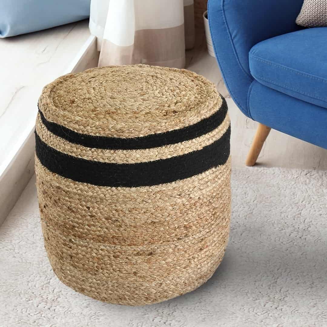 Placed next to a sofa on a carpet, the Jute Pouf with Black Accent, a braided and stitched ottoman made from jute & cotton, available at Sukham Home, a sustainable furniture, kitchen & dining and home decor store in Kolkata, India