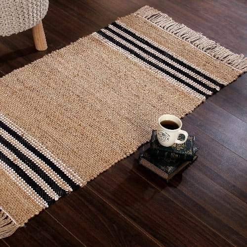 On a wood floor, the Handwoven Jute Rug with Black and White Stripes and Tassels, a small rectangle jute and black & white cotton carpet you can buy online at Sukham Home, a sustainable furniture, kitchen & dining and home decor store in Kolkata, India