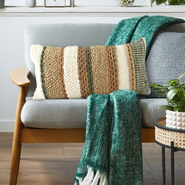 On a grey sofa with a green throw, the Boho Woven Cushion, a rectangle green, white and brown accent pillow available at Sukham Home, a sustainable furniture, kitchen & dining and home decor store in Kolkata, India