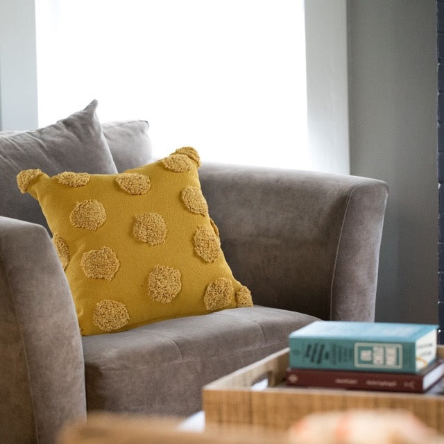 Placed on a grey sofa, the Gold Tufted Dots Cushion, a square yellow accent pillow available at Sukham Home, a sustainable furniture, kitchen & dining and home decor store in Kolkata, India