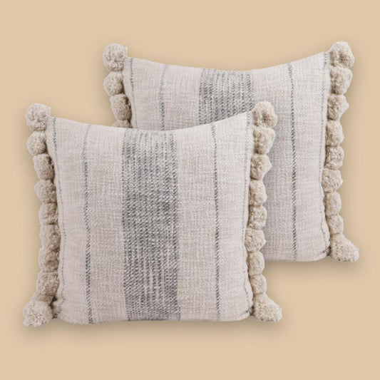 Against a yellow background, Graphite Cushions, a set of two white and grey linen accent pillows with pom poms you can buy online at Sukham Home, a sustainable furniture, kitchen & dining and home decor store in Kolkata, India