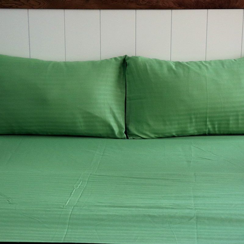 Placed on a bed, the two pillowcases and sheet from the Green Sateen Striped Bedsheet, a king size cotton bedsheet you can buy online at Sukham Home, a sustainable furniture, gardening and home decor store in Kolkata, India