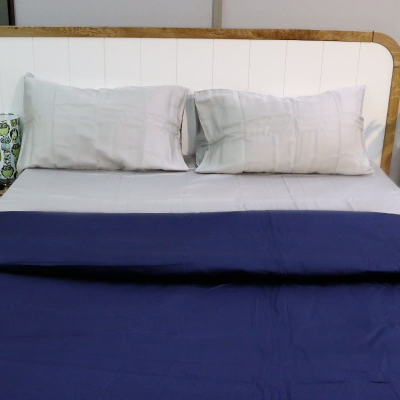 Placed on a bed with a blue duvet, the Silver Grey Classic Solid Bedsheet, a king size and queen size cotton bedsheet you can buy online at Sukham Home, a sustainable furniture, gardening and home decor store in Kolkata, India