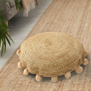 On a jute rug, the Jute Round Floor Cushion, a circular braided accent pillow with pom poms, available at Sukham Home, a sustainable furniture, kitchen & dining and home decor store in Kolkata, India