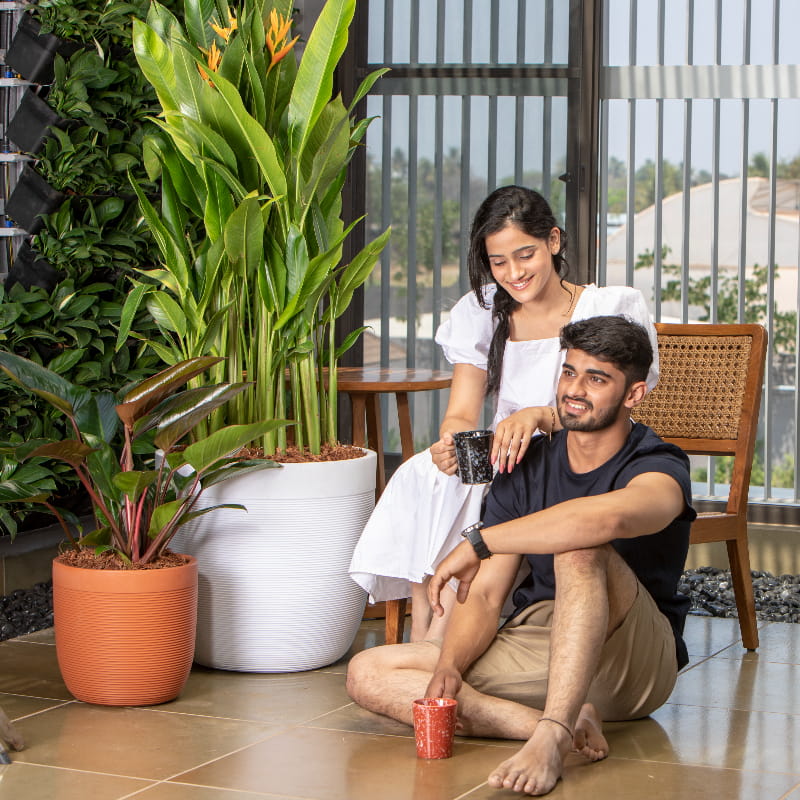 Placed outdoors with plants, the White Stone Milano, a round ribbed stone finish plastic planter that looks like granite available at Sukham Home, a sustainable furniture, gardening and home decor store in Kolkata, India