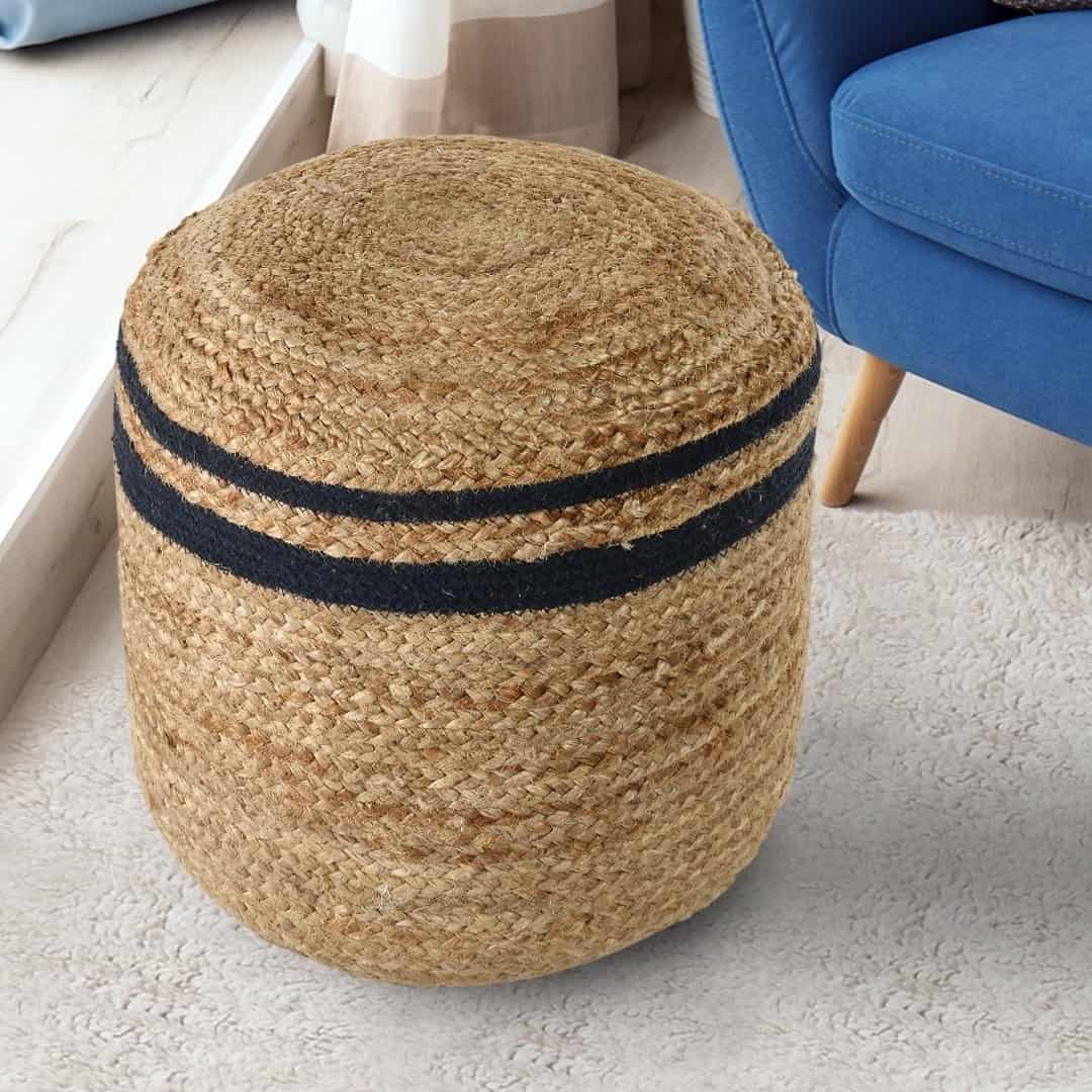 Placed next to a sofa on a carpet, the Jute Pouf with Navy Blue Accent, a braided and stitched ottoman made from jute & cotton, available at Sukham Home, a sustainable furniture, kitchen & dining and home decor store in Kolkata, India