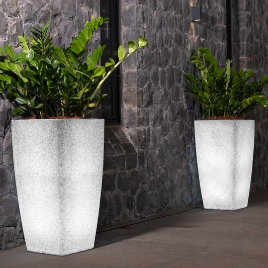 Placed against a stone wall, two Paris, a tall square stone finish plastic planter that looks like granite and comes with LED lights available at Sukham Home, a sustainable furniture, gardening and home decor store in Kolkata, India