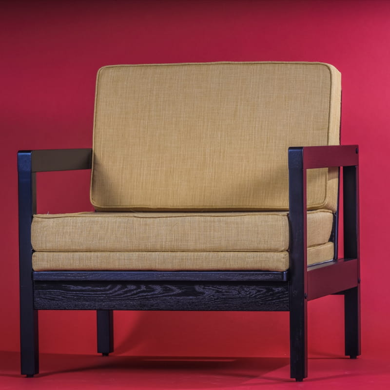 Against a red background, the Tan fabric in Ash Charcoal Sepia Armchair, a wooden cushioned midcentury seat you can buy online at Sukham Home, a sustainable furniture, kitchen & dining and home decor store in Kolkata, India