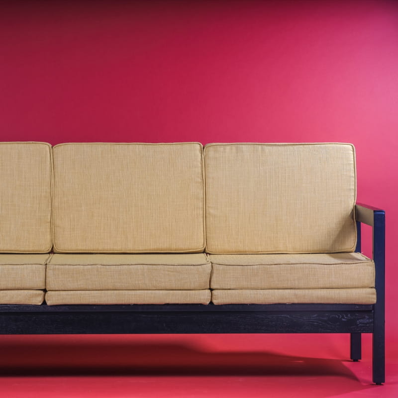 Against a red background, the Ash Charcoal Sepia Sofa, a wooden cushioned midcentury sofa you can buy online at Sukham Home, a sustainable furniture, kitchen & dining and home decor store in Kolkata, India